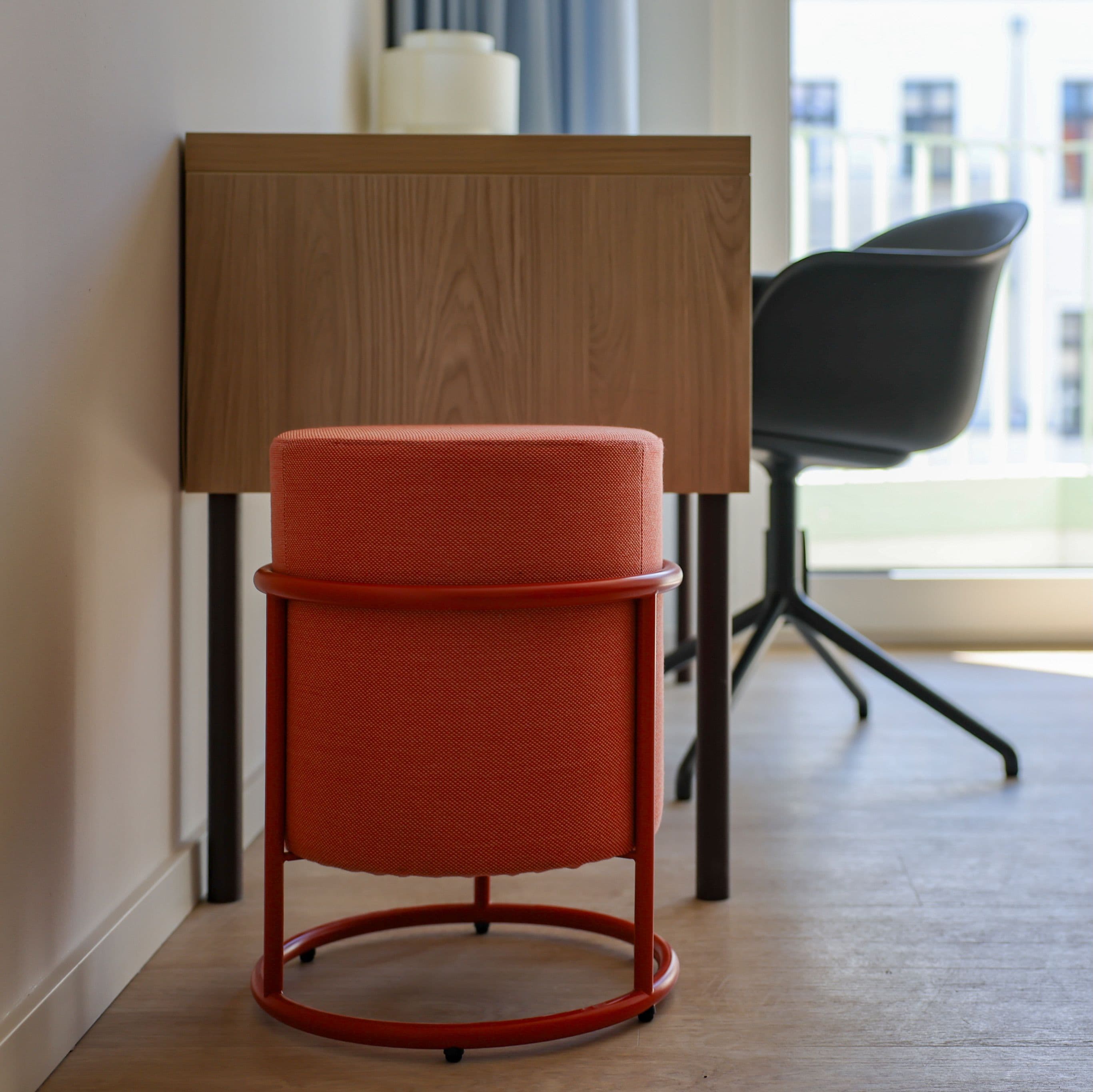 Einrichtungsdetail – runder Polsterhocker in Orangerot mit rotem Metallgestell vor einem Schreibtisch aus Holz, dahinter ein schwarzer Schalenstuhl am Fenster