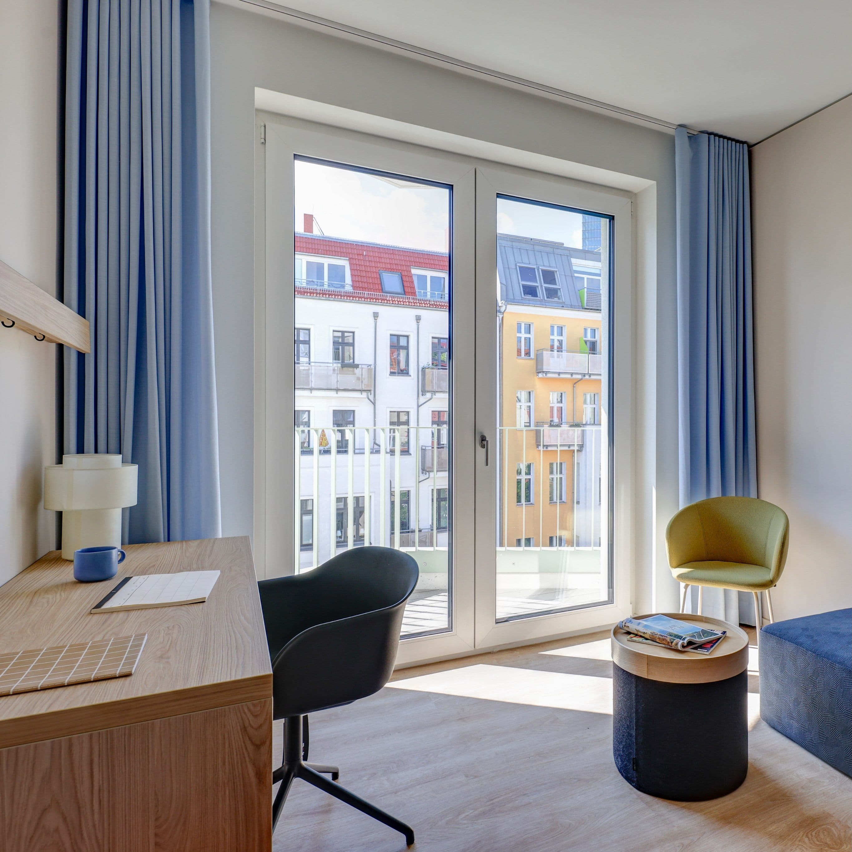 Premium Apartment – Holzschreibtisch mit schwarzem Drehstuhl vor bodentiefen Balkontüren mit Blick auf Altbaufassaden, dazu hellblaue Vorhänge, gelbgrüner Sessel und blaue Polsterbank