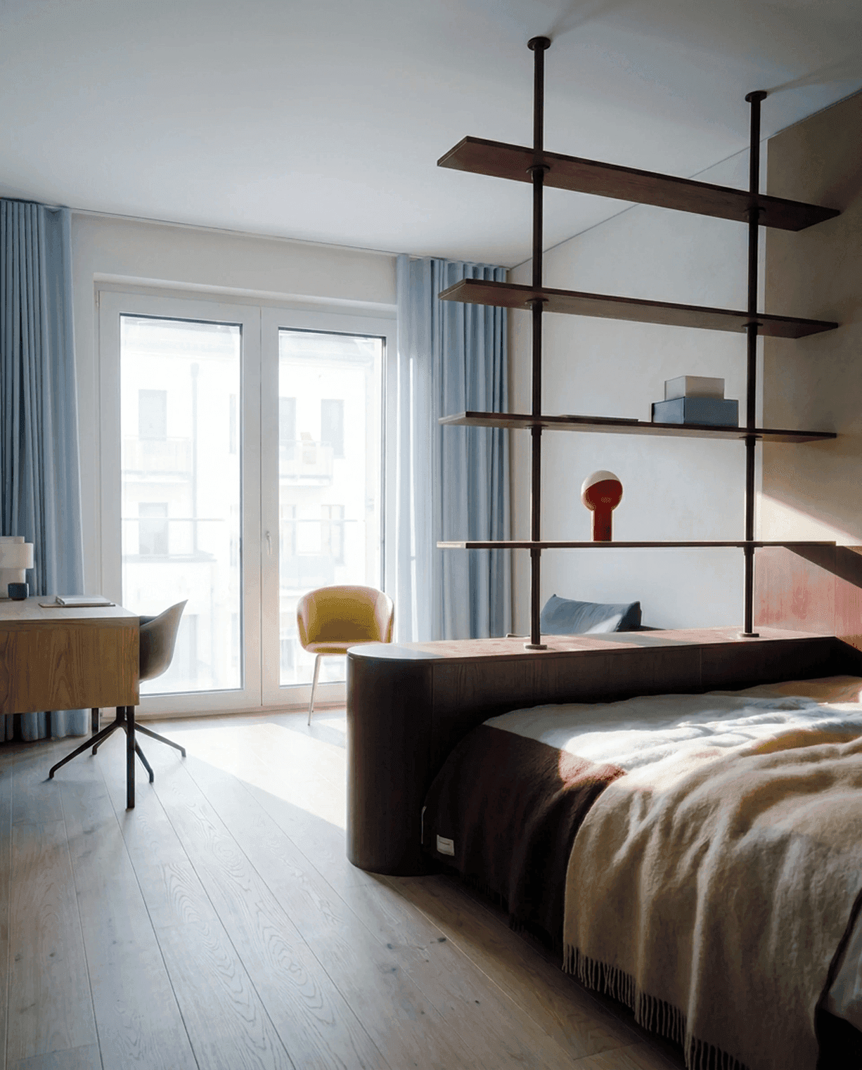 Premium Apartment Innenansicht: Doppelbett mit dunklem gerundetem Kopfteil und Regal-Raumteiler, Schreibtisch und gelber Loungesessel an der bodentiefen Balkontür mit hellblauen Vorhängen im Morgenlicht.