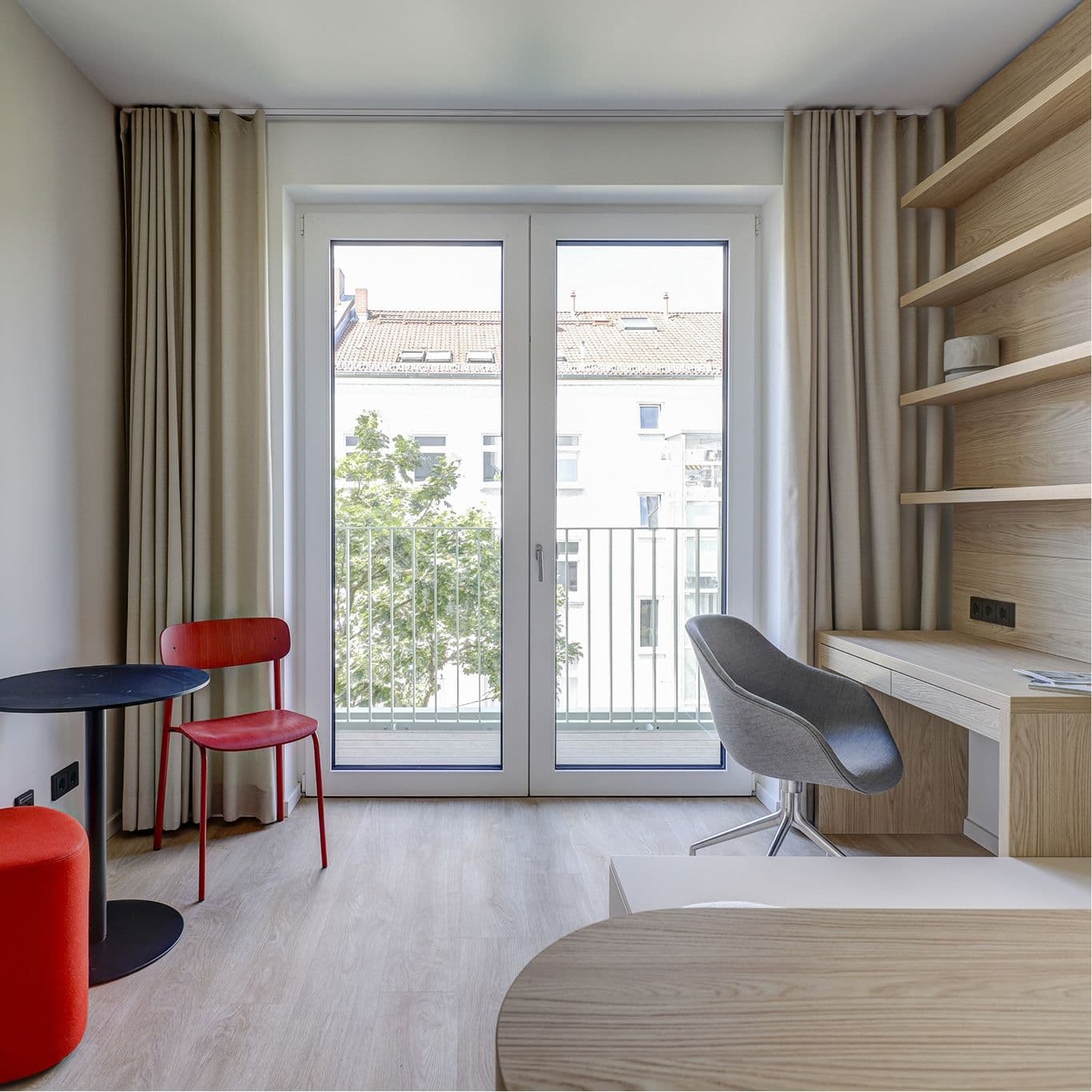 Studio Compact – helles Apartment mit bodentiefen Fenstertüren zum französischen Balkon, kleinem schwarzem Tisch mit rotem Stuhl und Hocker sowie Schreibtisch und Wandregal aus hellem Holz