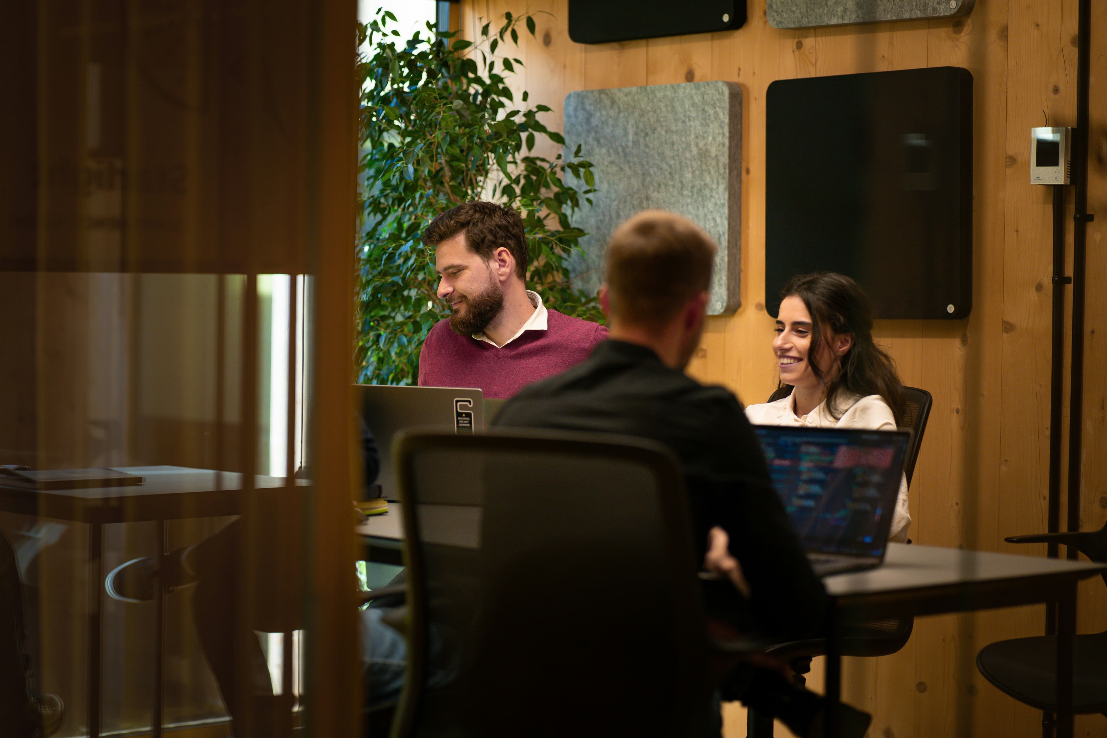 Coworking-Raum mit Holzvertäfelung und Akustikpaneelen: drei Personen arbeiten lächelnd an Laptops, im Hintergrund eine große Grünpflanze.