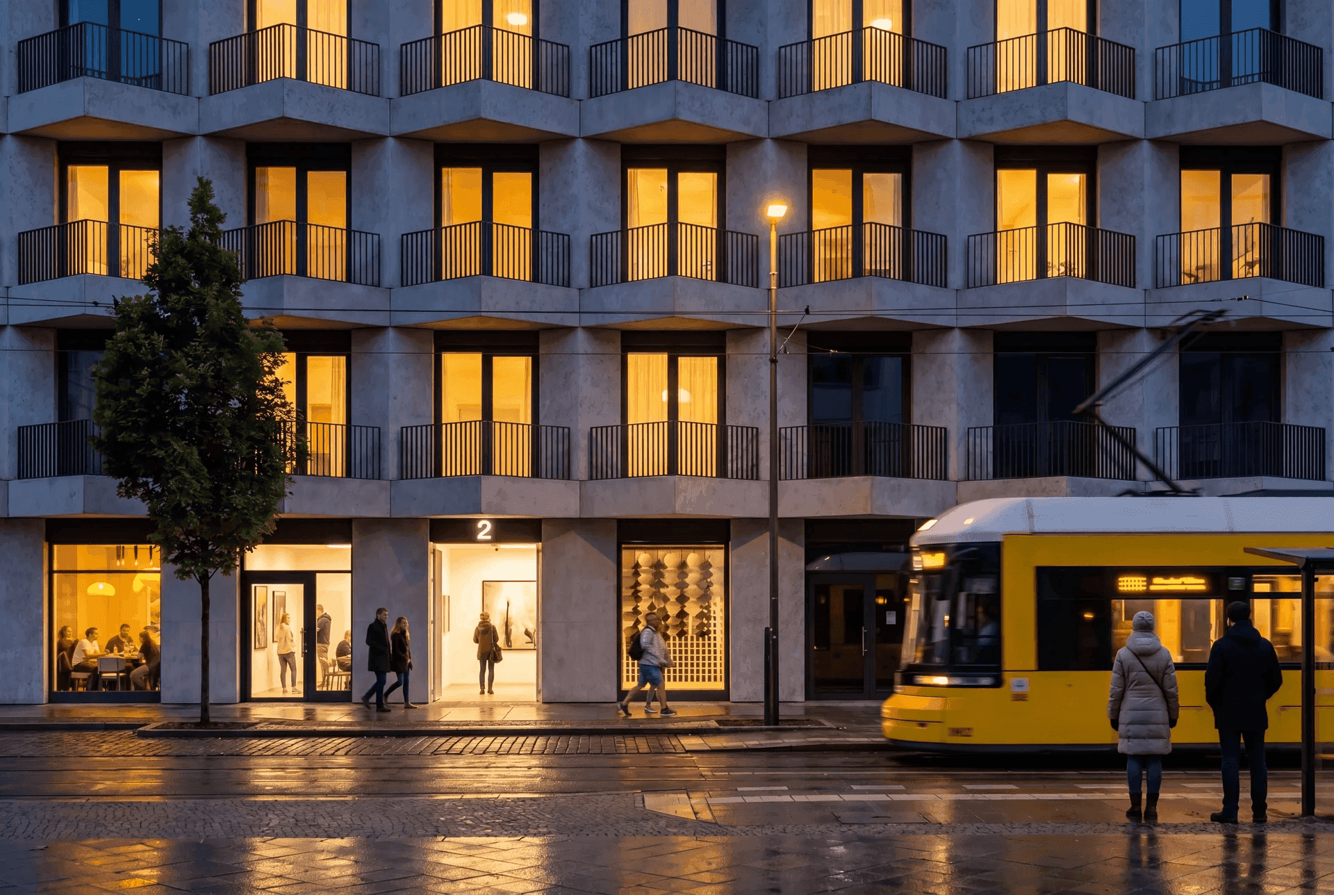 Friedrich-Fassade in der Abenddämmerung: warm erleuchtete Fenster mit Balkonen, eine gelbe Berliner Tram fährt vorbei, belebtes Erdgeschoss mit Galerie und Café, nasses Pflaster spiegelt die Lichter.