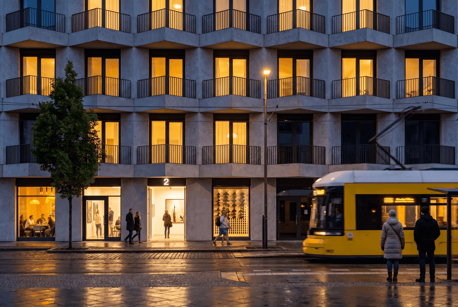 Friedrich-Fassade in der Abenddämmerung: warm erleuchtete Fenster mit Balkonen, eine gelbe Berliner Tram fährt vorbei, belebtes Erdgeschoss mit Galerie und Café, nasses Pflaster spiegelt die Lichter.