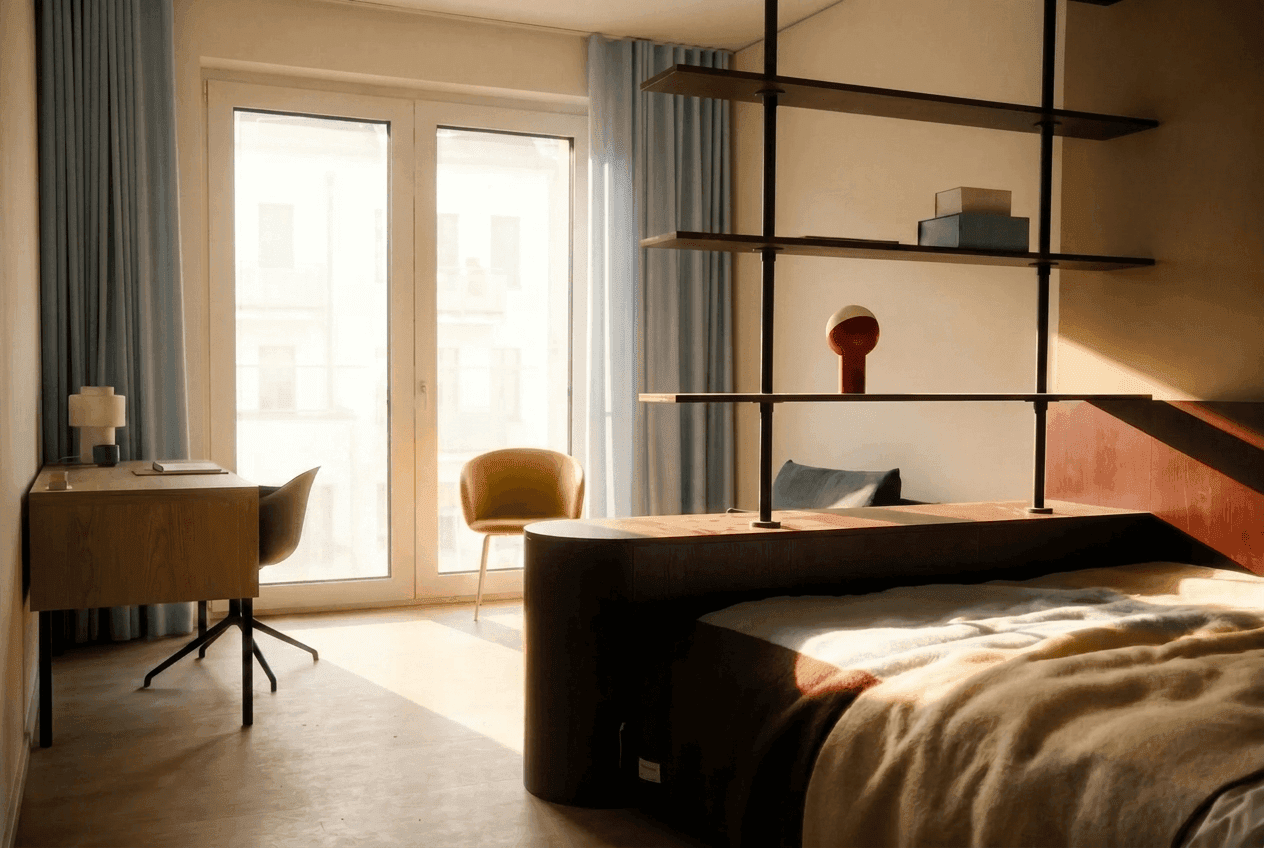 Friedrich Premium Apartment Schlafzimmer: dunkelbrauner gerundeter Bettrahmen mit Regaltrennwand, Schreibtisch und gelber Lounge-Sessel, Balkonflügeltüren mit Stadtblick im Morgenlicht
