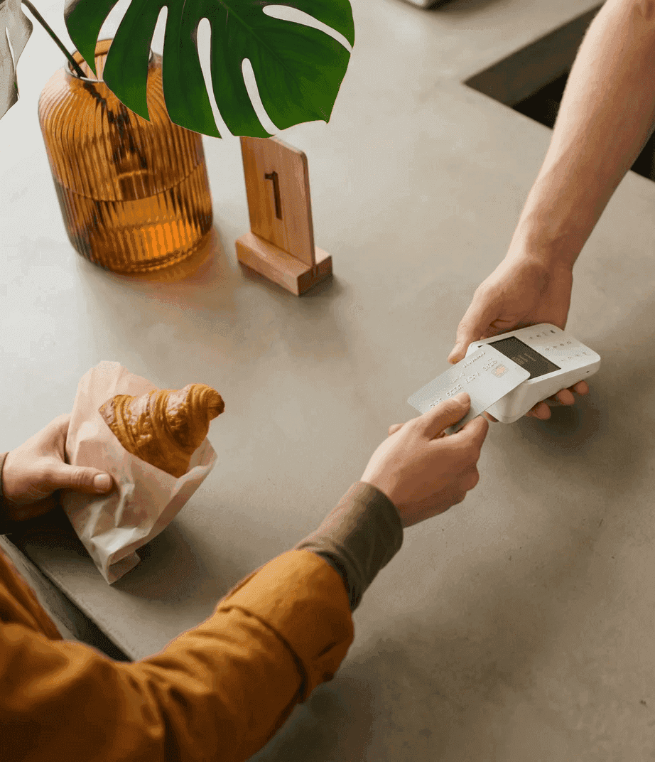 Kaffeebar von oben: Person zahlt kontaktlos per Kreditkarte am Terminal, daneben ein Croissant in Pergaminpapier und eine Monstera-Pflanze in einer orangefarbenen Vase