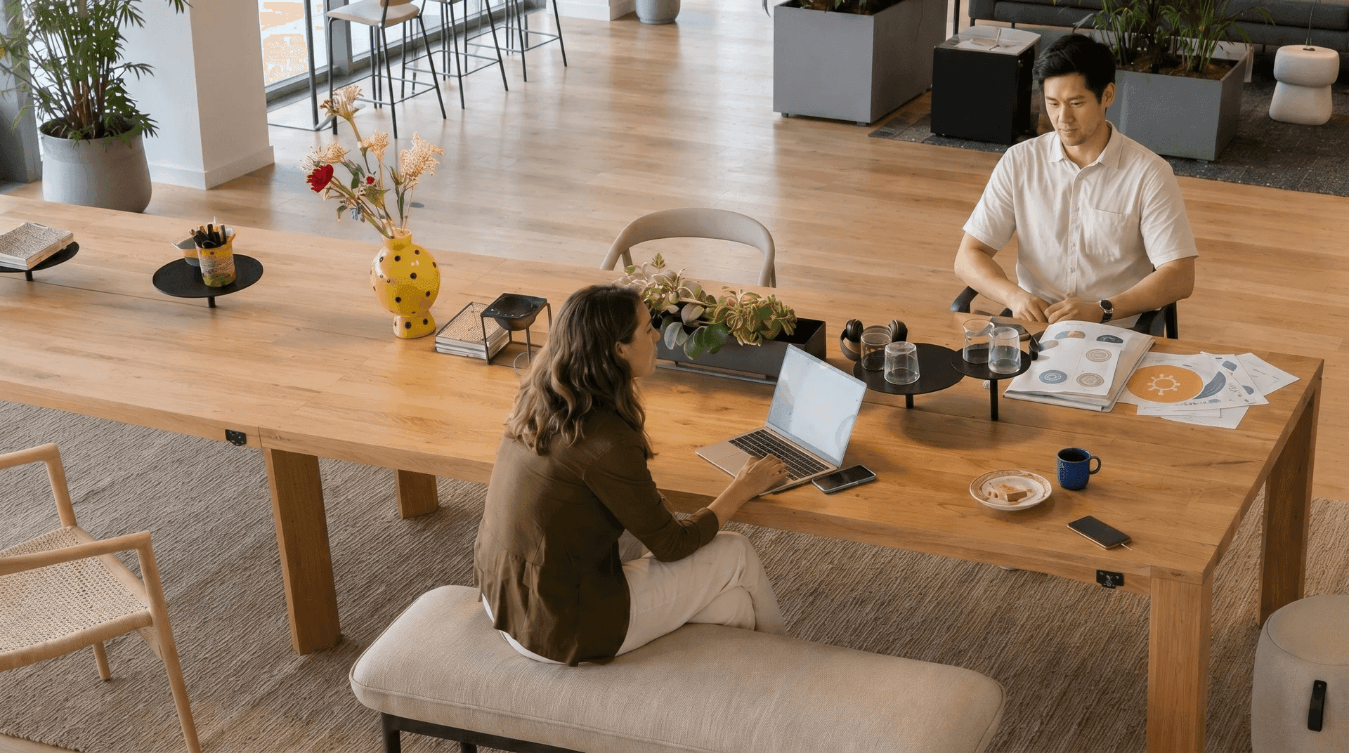 Coworking-Bereich von oben: eine Frau mit Laptop auf einer gepolsterten Sitzbank und ein Mann mit Unterlagen sitzen an einem langen Holztisch, umgeben von Grünpflanzen und Lounge-Möbeln.