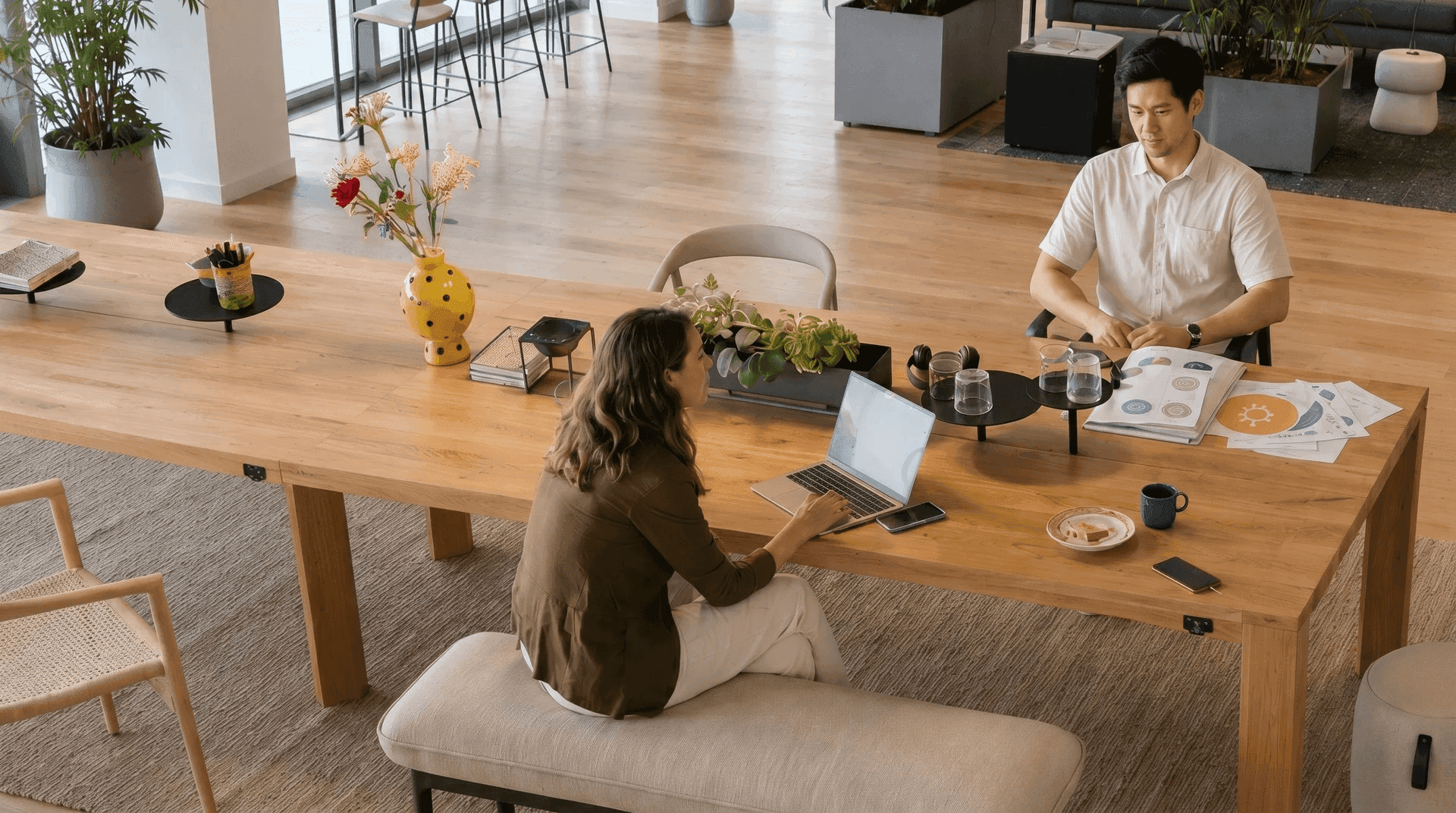 Coworking-Bereich von oben: eine Frau mit Laptop auf einer gepolsterten Sitzbank und ein Mann mit Unterlagen sitzen an einem langen Holztisch, umgeben von Grünpflanzen und Lounge-Möbeln.