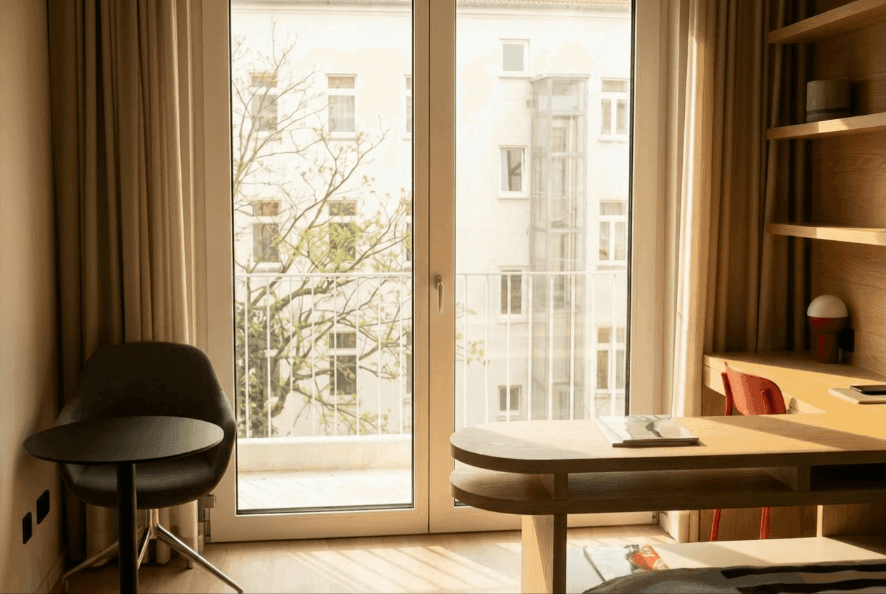 Sonnendurchflutete Apartment-Ecke: bodentiefe Balkontür mit Blick auf Berliner Altbauten und Baumkronen, dunkler Schalensessel, runder Beistelltisch und Holzschreibtisch mit Regalwand.