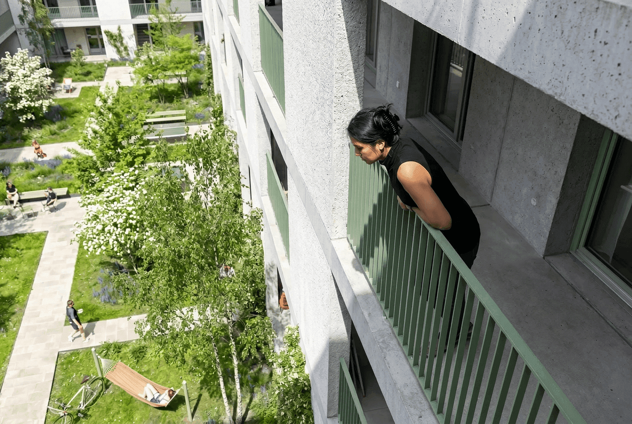 Blick vom Balkon auf den begrünten Friedrich-Innenhof: eine Frau lehnt am grünen Geländer, darunter Birken, blühende Beete, eine Hängematte und entspannende Bewohner.