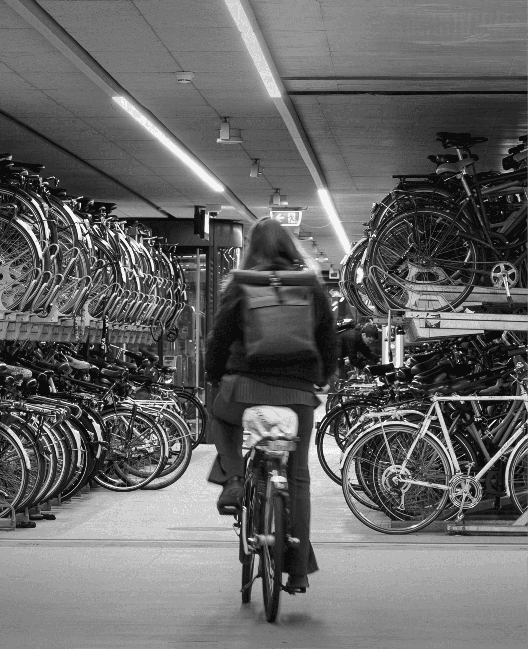 Schwarz-Weiß-Aufnahme einer Radfahrerin mit Rucksack in einer großen unterirdischen Fahrradabstellanlage